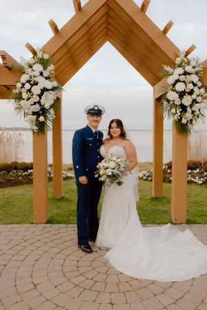 Coast Guard groom in dress uniform poses with bride holding bouquet under wooden wedding arch.