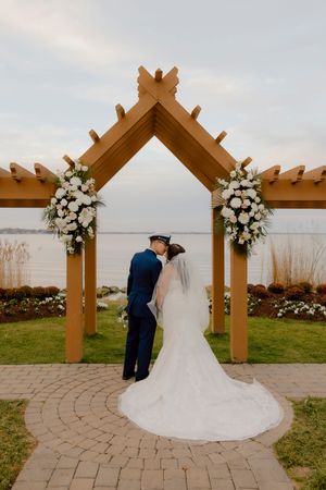 Bride and groom in military uniform embrace under wooden arch with waterfront backdrop.
