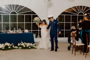 Bride and groom in military dress uniform enter wedding reception tent holding hands.