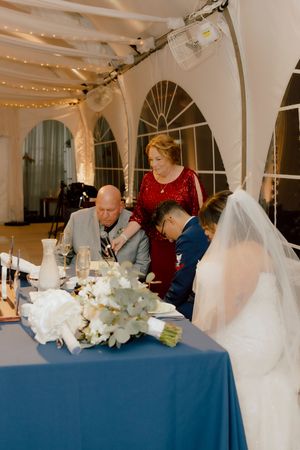 Wedding party signs marriage documents at blue-draped table inside elegant tented venue.