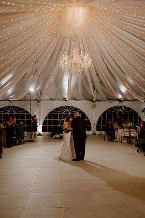 Bride dances with father under chandelier in draped white tented wedding reception venue.