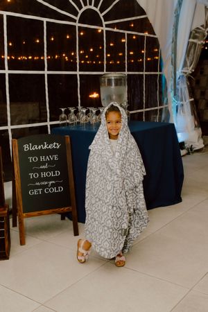Smiling young girl wrapped in gray patterned blanket at wedding reception near blanket station.