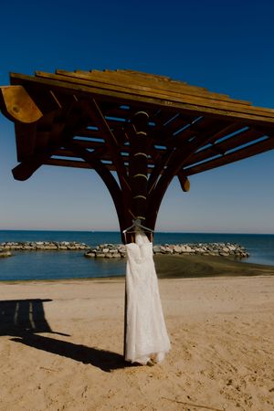 White wedding dress hanging on wooden beach gazebo with ocean and rocky shoreline in background.