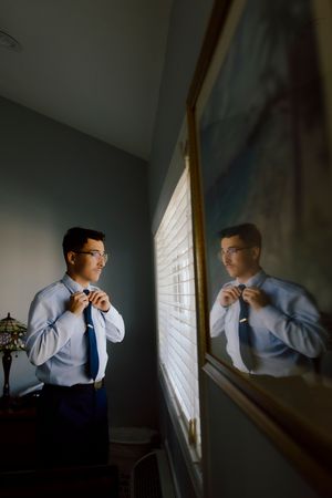 Groom adjusting tie while looking at reflection in mirror in dimly lit room.