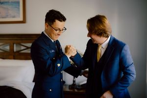 Groom in military uniform adjusts cufflinks with groomsman in blue suit before wedding ceremony.