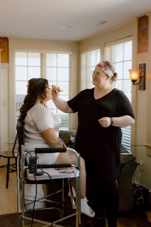 Makeup artist applying makeup to bride seated in chair near bright windows indoors.