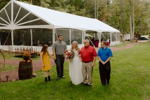 A smiling bride in white holds a bouquet while walking with family members toward a wedding tent.