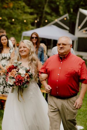 A radiant bride carrying a fall bouquet walks down the aisle with her father in a red shirt.