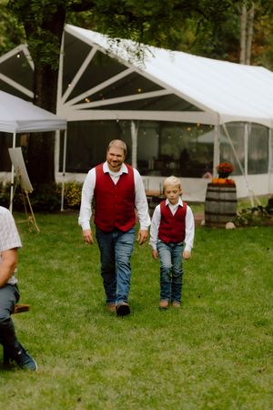 A man and young boy in matching red vests and jeans walk together across a lawn at a wedding.
