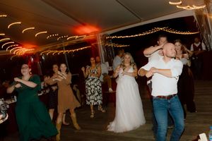 Wedding guests dance joyfully with the bride in a white gown under string lights at a reception.