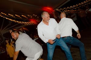 Three men in white shirts dance energetically together under string lights at a wedding reception.