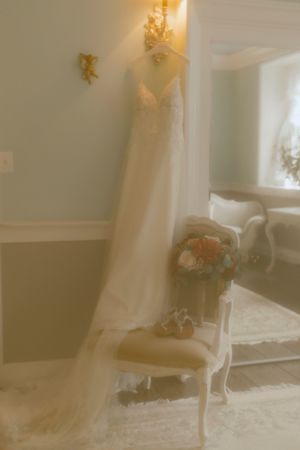 Elegant wedding gown hanging in a softly lit room beside a floral bouquet resting on a vintage chair.