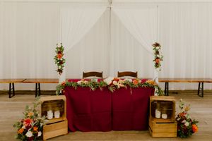 Wedding sweetheart table draped in burgundy linen, adorned with lush floral garlands and candles in a white tent venue.