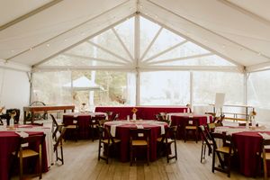 Elegant wedding reception tent with burgundy tablecloths, wooden chairs, and large windows overlooking lush greenery.