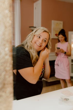 Smiling blonde bride-to-be in black shirt getting ready, reflected in mirror before her wedding.