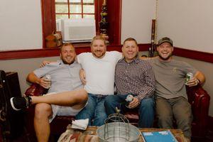 Four smiling groomsmen sit together on a couch posing for a photo before the wedding celebration.