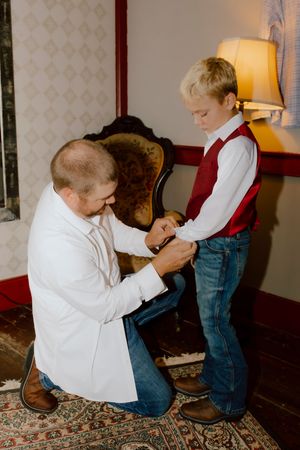 A father kneels to help his young son in a red vest and jeans with cufflinks before a wedding ceremony.