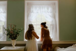 A bride in a white gown and bridesmaid look out a window together during wedding preparations.