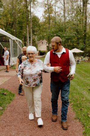 Young man in red vest escorts elderly woman along a garden path at an outdoor wedding reception.