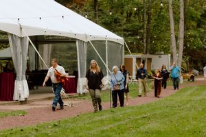 Guests walk along a path toward a white tent at an outdoor wedding venue surrounded by trees.