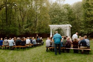 Wedding guests seated on wooden benches at an outdoor ceremony facing a white pergola altar.