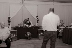 Black and white wedding reception photo with guests seated as a man delivers a heartfelt speech to the newlyweds.
