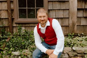 Groom in red vest and white shirt smiling while crouching near stone wall with ivy backdrop.