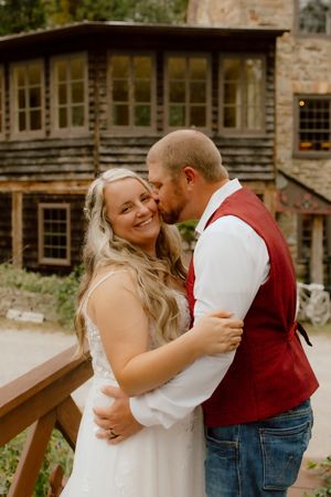 Groom in red vest kissing bride on cheek as she smiles in white wedding gown outdoors.