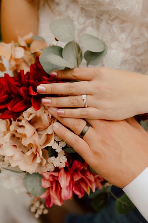 Bride and groom displaying wedding rings over colorful bridal bouquet with red and blush blooms.
