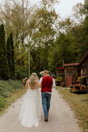 Bride and groom walking hand in hand down gravel path surrounded by autumn trees.
