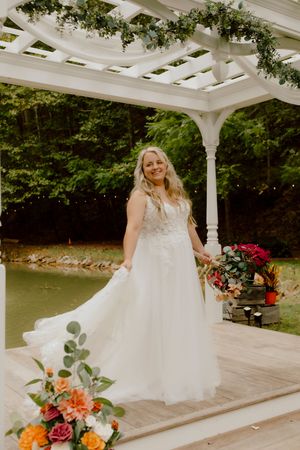 Smiling bride in white lace ball gown holding bouquet under white pergola with greenery near pond.