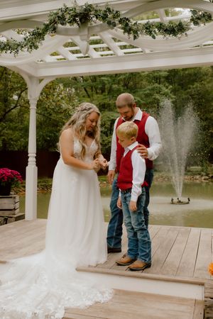 Bride and groom share a moment with a young boy in a red vest under a white pergola by a fountain.