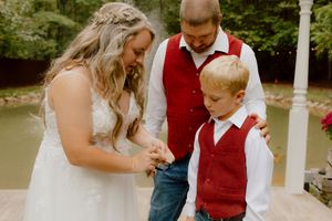 Bride, groom in red vest, and young boy share a tender moment together during outdoor wedding ceremony.