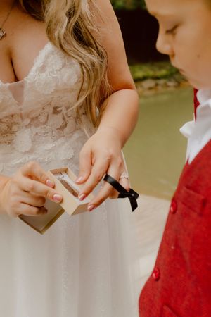 Bride in lace wedding gown opens a ring box with a young boy in a red vest beside her.