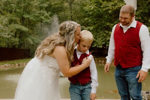 Bride kisses young boy in red vest on the cheek while groom smiles during outdoor wedding ceremony.