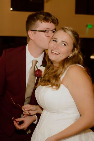 Groom in burgundy suit kissing smiling bride on cheek during wedding reception.