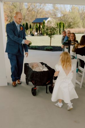Groomsman in blue suit pulling wagon with baby as flower girl walks down aisle.