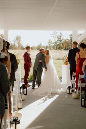 Bride and groom sharing first kiss at outdoor fall wedding ceremony with guests watching.