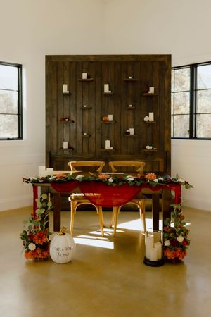 Rustic sweetheart table with red tablecloth, floral garland, and candle wall backdrop at wedding reception.