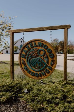 Wooden Hazelwood Weddings sign hanging on post outside white barn wedding venue on sunny day.