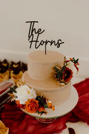 Two-tier rustic wedding cake with fall florals and The Horns cake topper on a red tablecloth.