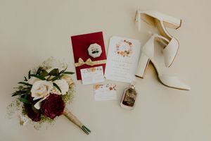 Flat lay of wedding details including white heels, bouquet, invitation suite, and perfume bottle.