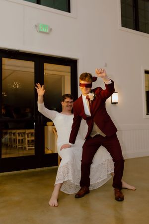 Groom blindfolded in burgundy suit dancing energetically with guest at wedding reception.
