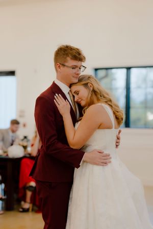Bride and groom sharing intimate first dance, groom in burgundy suit and glasses.