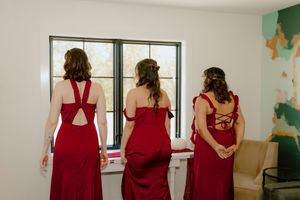 Three bridesmaids in red dresses standing together looking out a window before the wedding.