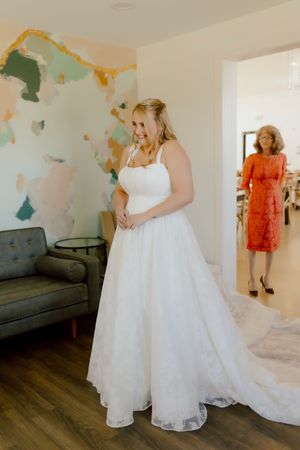 Smiling bride in white ballgown wedding dress with mother watching proudly in doorway.