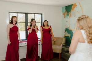 Bridesmaids in red dresses reacting emotionally to first look at bride in white wedding gown.