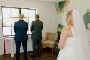 Bride in white gown and veil approaches two men standing by a window indoors.