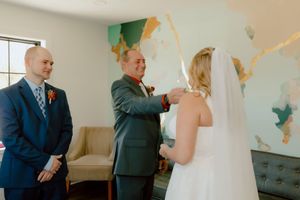 Father adjusts bride's veil while groom watches during an intimate wedding preparation moment.
