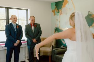 Bride in white wedding dress and veil reveals herself to two smiling men indoors.
